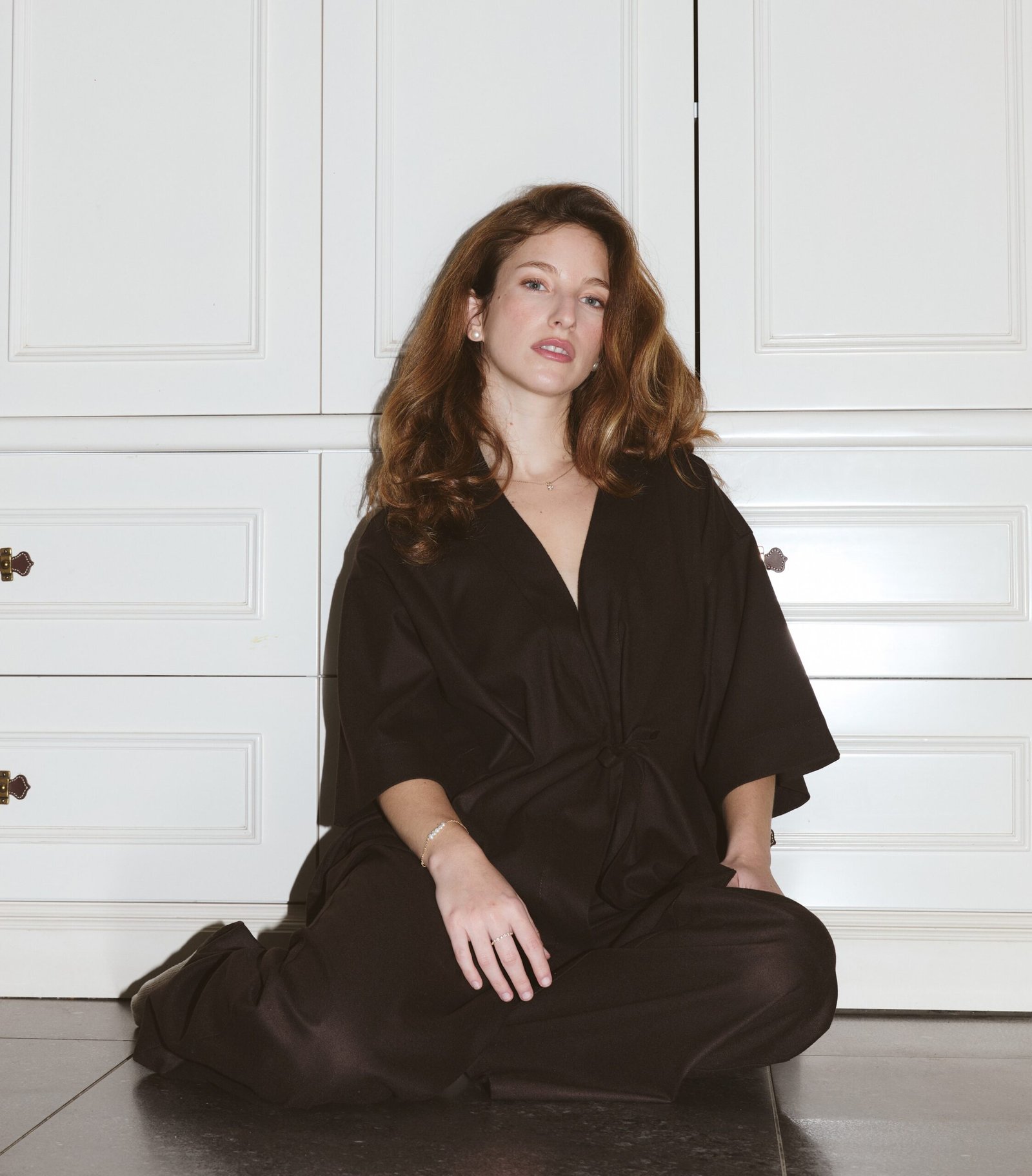 הדוגמנית יושבת על רצפה בצבע חום כאשר מאחוריה ארון לבן והיא לובשת את סט הבגדים l'amandier